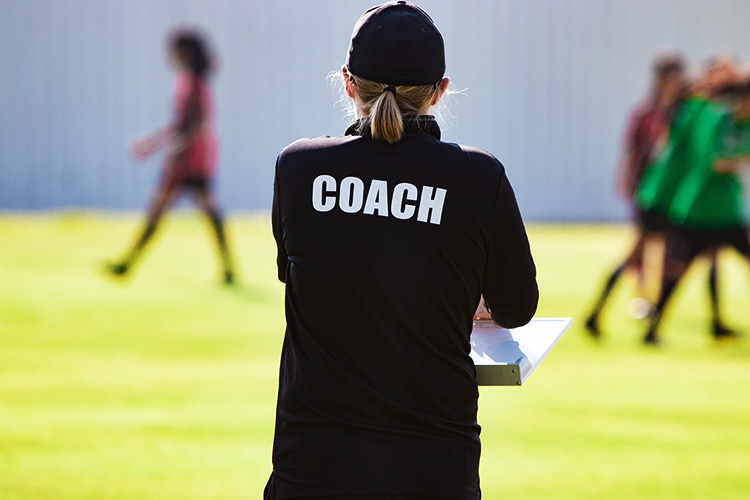 Female soccer coach on pitch watching and assessing teenage women participating in a tryout to be evaluated for their talent level to play professional or collegiate soccer.