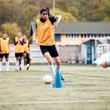 Female soccer player leading ball among cones during sports training.