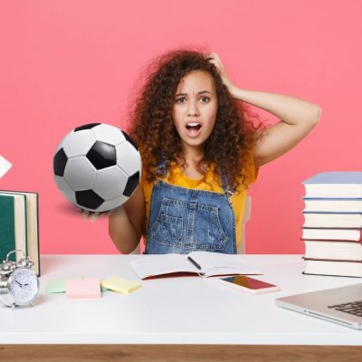 Young girl looking stressed at a desk holding ball in right hand and books and laptop for homework on the desk