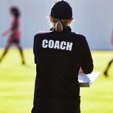 Female soccer coach on pitch watching and assessing teenage women participating in a tryout to be evaluated for their talent level to play professional or collegiate soccer.