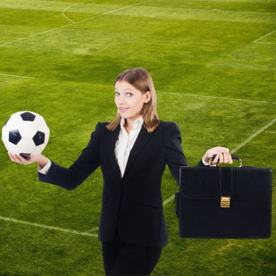 Women on soccer field wearing black suit holding a soccer ball in right hand and briefcase in her lefthand.