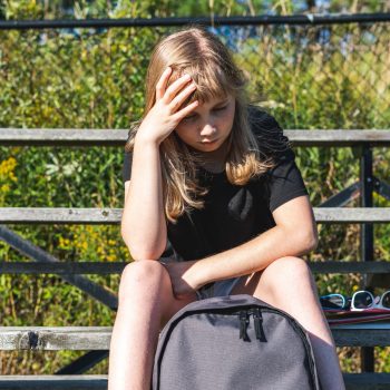 Young high school soccer player stressed sitting on bleacher by side of soccer field with hand on her head, backpack in front of her.