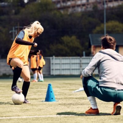 Female soccer player dribbling around cones on field with coach looking on.